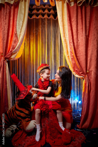 Family during a stylized theatrical circus photo shoot in a beautiful red location. Models mother and son posing on stage with circus curtain