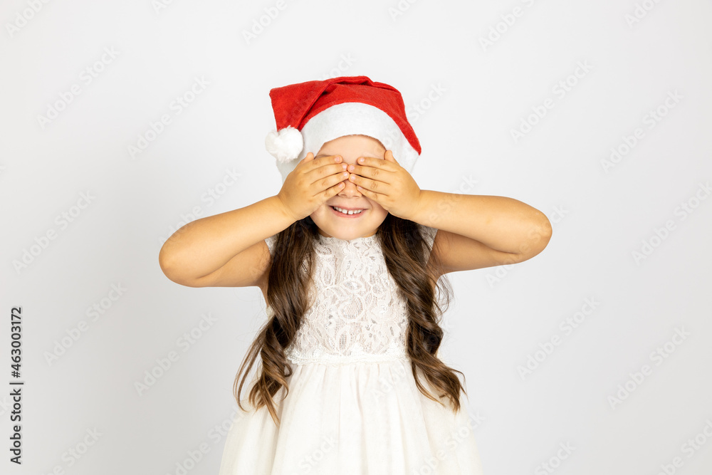 portrait of joyful girl with long hair in white dress, in red Santa Claus hat covering eyes with palms, isolated on white background, concept of waiting for New Year gift. 