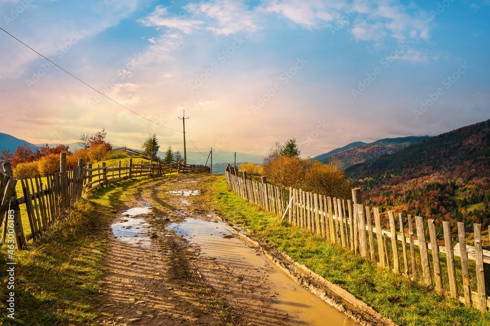 A path along the ridge of a hill with a large puddle and an old fence against the backdrop of a colorful forest A path along the ridge of a hill with a large puddle and an old fence against the backdrop of a colorful forest
