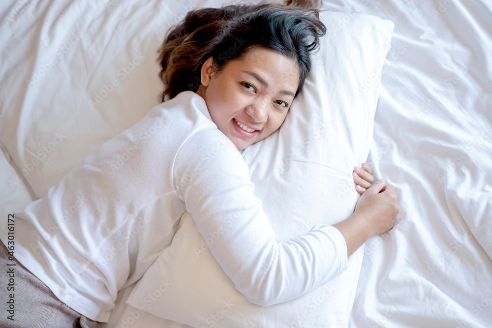 A young Asian woman is comfortable in bed, hugging a soft pillow in a hotel room with a bright, smiling, happy face. It is a good rest on a long vacation. Top view A young Asian woman is comfortable in bed, hugging a soft pillow in a hotel room with a bright, smiling, happy face. It is a good rest on a long vacation. Top view