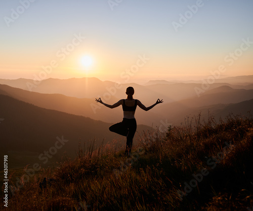 Back view of slim woman performing yoga pose on grassy hill with orange sky on background. Fit woman standing on one leg and doing yoga exercise outdoors at sunset.