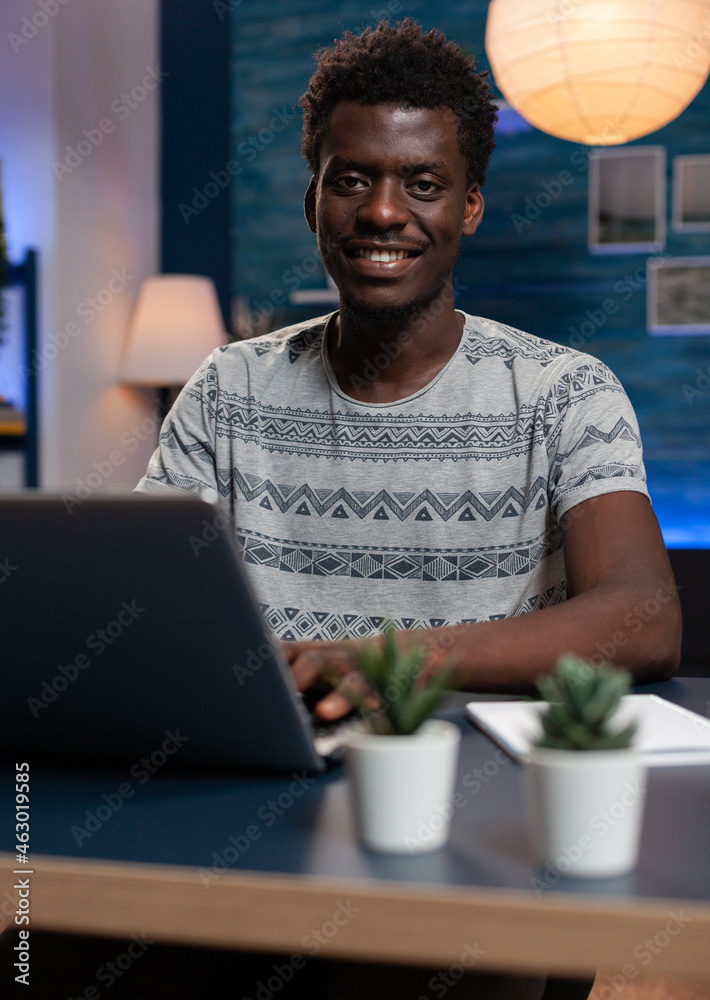 Portrait of smiling african american entrepreneur man browsing management information on laptop computer. Young freelancer working remote at online project in living room late at night Portrait of smiling african american entrepreneur man browsing management information on laptop computer. Young freelancer working remote at online project in living room late at night