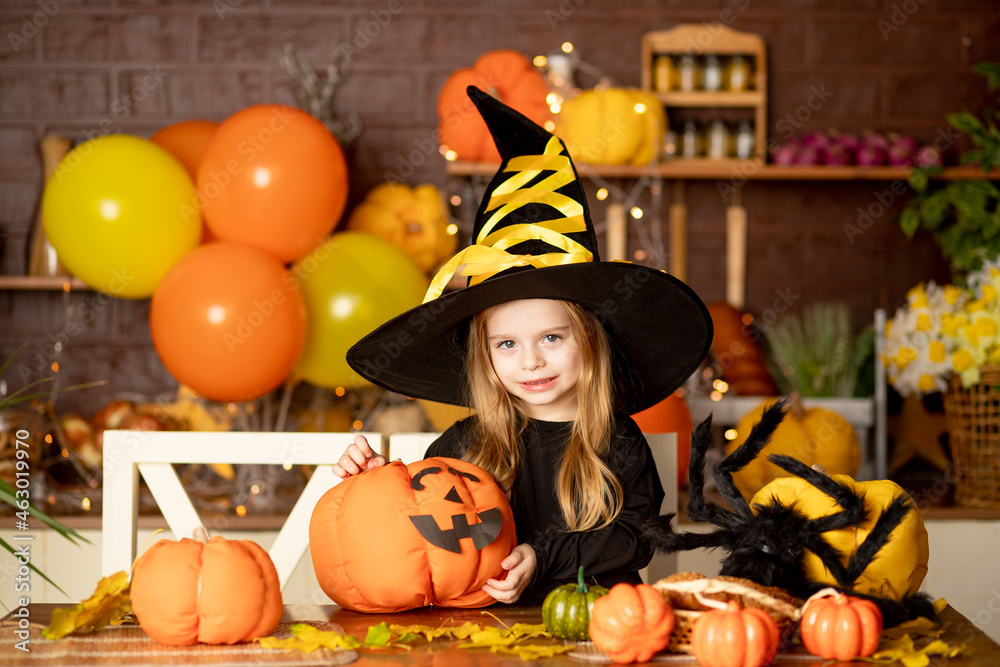 halloween, child a girl in a witch costume with pumpkins and a big spider in a dark kitchen decorates a pumpkin during the Halloween celebration