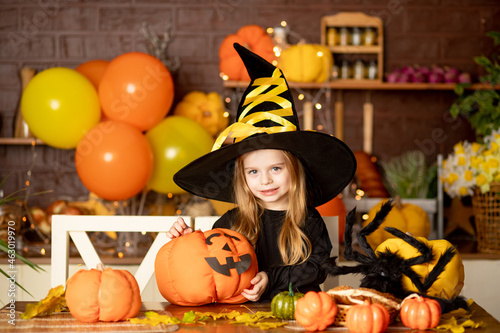 halloween, child a girl in a witch costume with pumpkins and a big spider in a dark kitchen decorates a pumpkin during the Halloween celebration