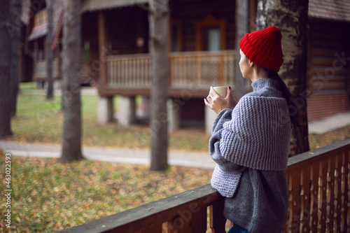 woman in a red hat and scarf and a mug stands at a wooden house in the woods