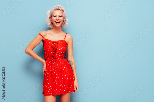 Portrait of young beautiful smiling female in trendy summer red dress. Sexy carefree woman posing near blue wall in studio. Positive blond model having fun and going crazy. Cheerful and happy