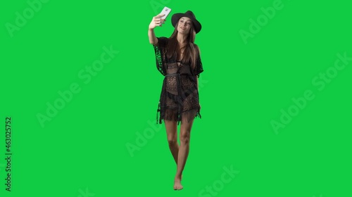 Pretty influencer summer girl in tunic walking and vlogging with smart phone. Full body on green screen chroma key background