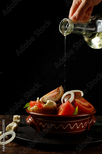 Homemade salad of different varieties of tomatoes in a clay bowl. And the hand pours oil from a glass jug. Low key, Vertical, Selective focus
