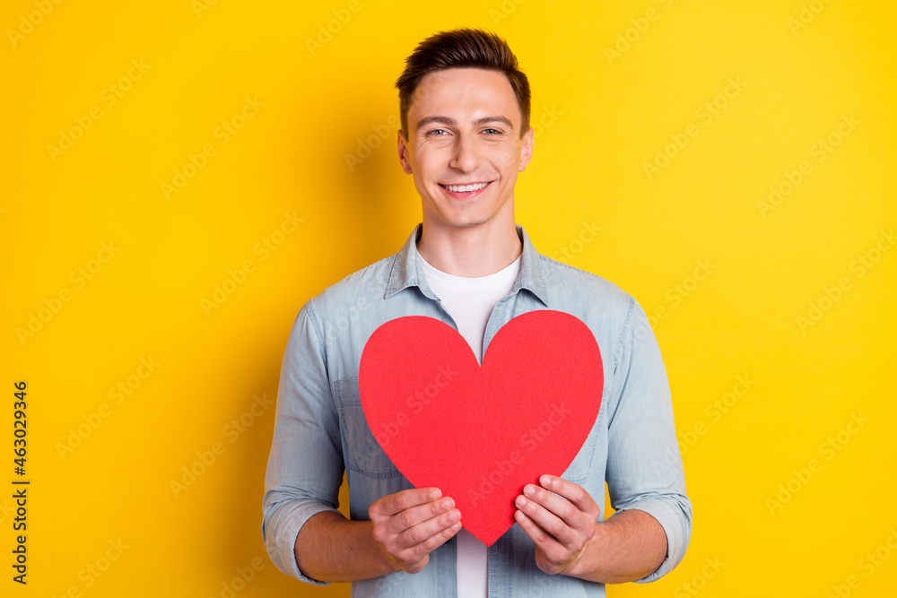 Portrait of attractive cheerful guy holding in hands big heart card isolated over bright yellow color background Portrait of attractive cheerful guy holding in hands big heart card isolated over bright yellow color background