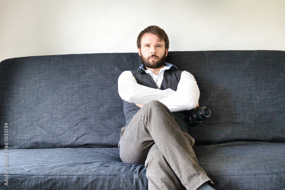 A bearded photographer is sitting on a couch and posing with his digital camera. He is wearing a grey vest and a white shirt. A bearded photographer is sitting on a couch and posing with his digital camera. He is wearing a grey vest and a white shirt.