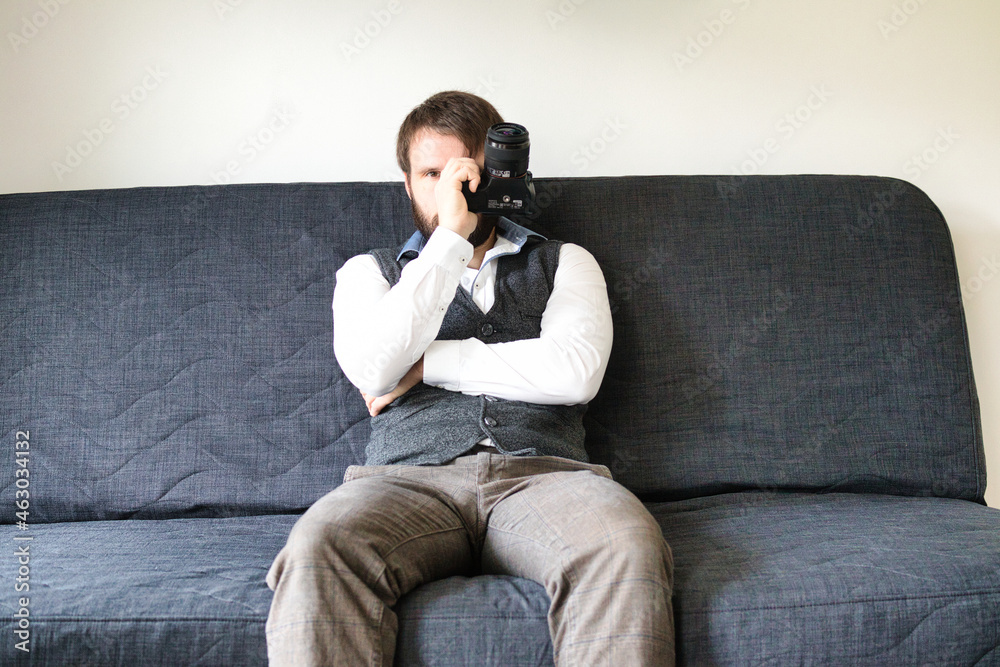 A bearded photographer is sitting on a couch and posing with his digital camera. He is wearing a grey vest and a white shirt. A bearded photographer is sitting on a couch and posing with his digital camera. He is wearing a grey vest and a white shirt.