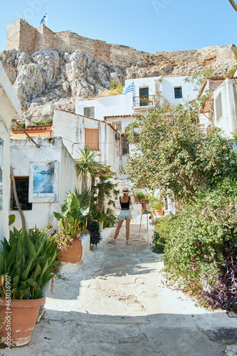 Narrow greek street with many plants and houses on the mountain and a girl in a hat. Passion for travel and tourism. Back view of the girl.