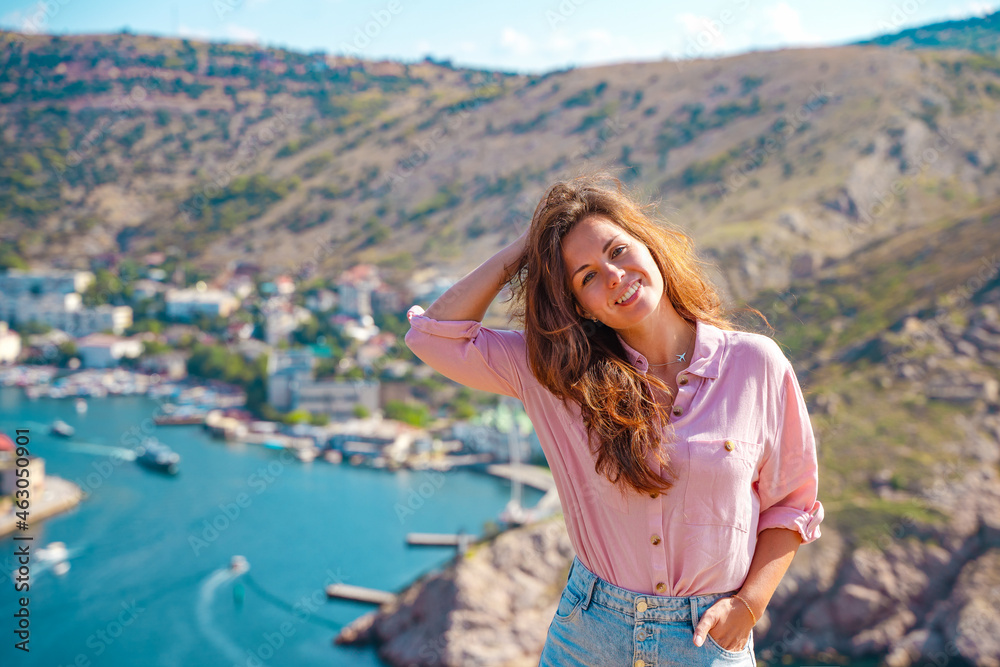 A young beautiful woman admires the picturesque landscape with a view of Balaclava with yachts and a colorful bay in summer. Postcard view of the tourist Crimea.