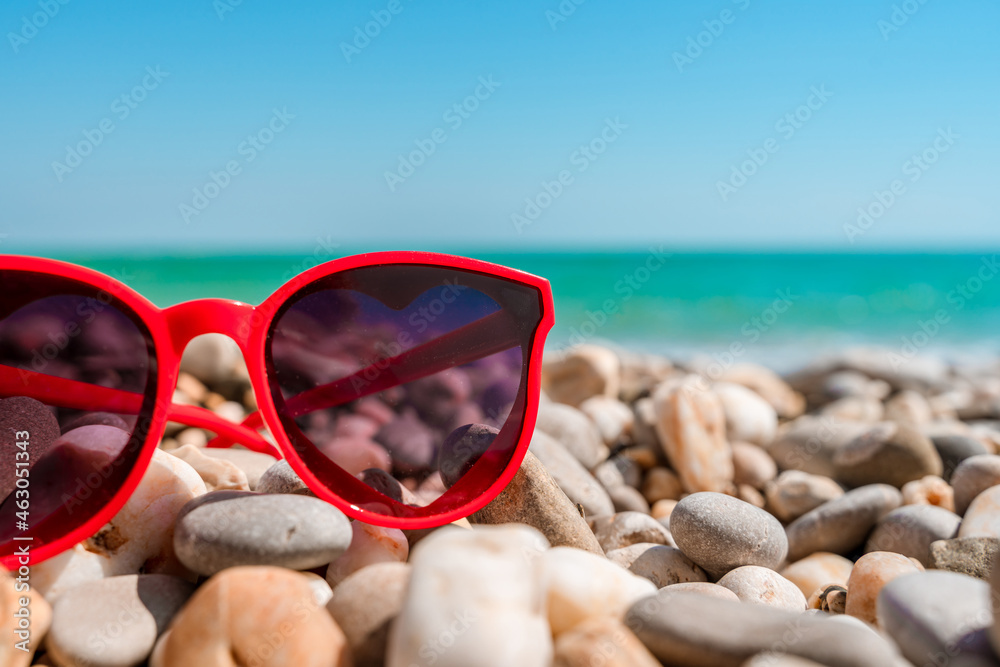 Red sunglasses in the form of hearts lie on pebbles on the beach in front of the azure sea. The concept of a relaxing vacation. Beautiful banner for travel