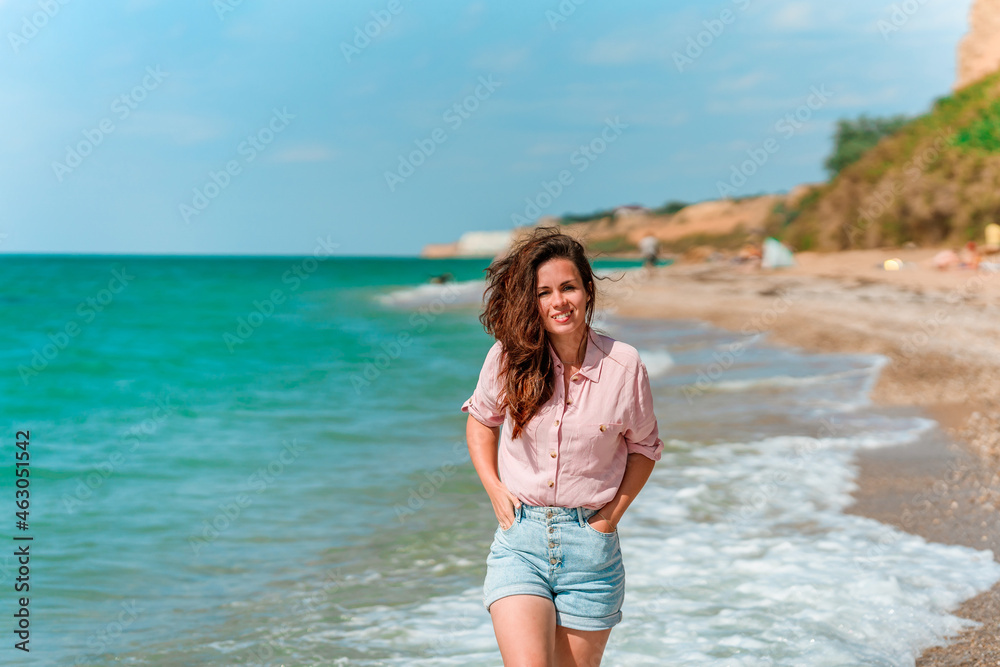 A charming young woman with long hair and wearing a shirt walks along the beach on a sunny day along the azure sea or ocean. The concept of summer holidays A charming young woman with long hair and wearing a shirt walks along the beach on a sunny day along the azure sea or ocean. The concept of summer holidays