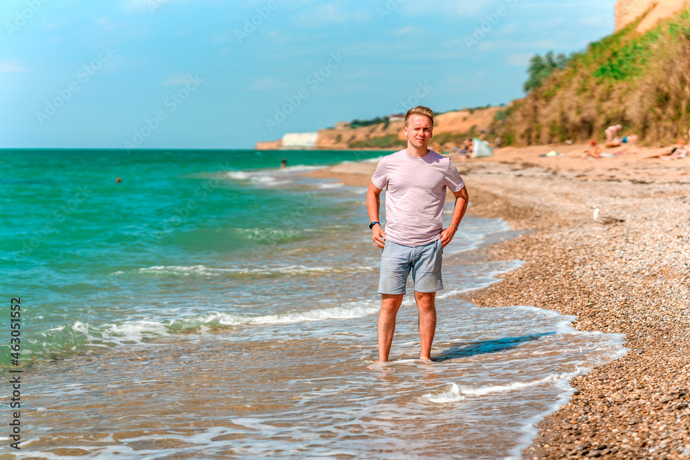 A young blond man walks on the beach near the azure ocean or sea. The concept of a happy summer vacation