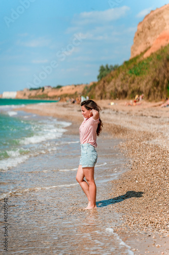 A charming young woman with long hair and wearing a shirt walks along the beach on a sunny day along the azure sea or ocean. The concept of summer holidays