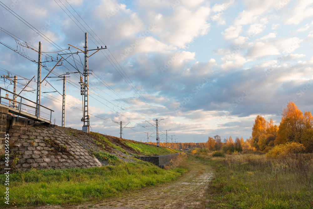 Landscape with green grass, yellow-leaved trees, footpath and and railway mound with poles and electric wires under blue sky with cumulus clouds