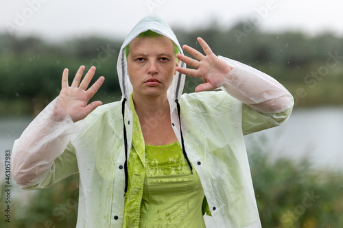 woman in bright clothes with green hair in transparent raincoat stands on bridge over river in village