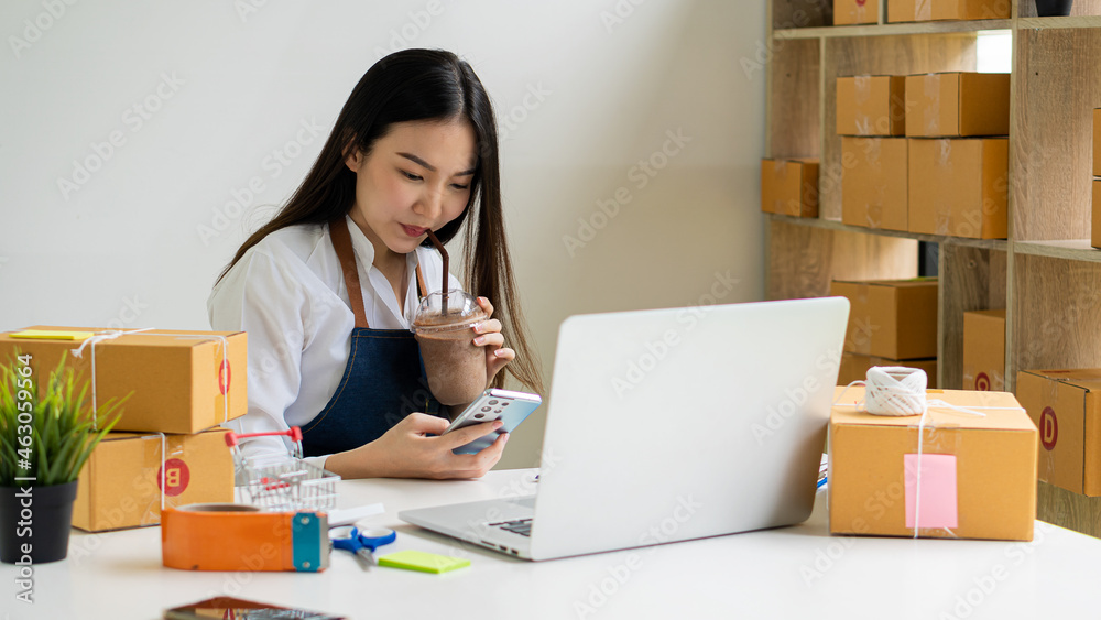 Beautiful Asian freelance SME online shopping business working on laptop computer with parcel box at home desk. delivery concept young woman owning a small business online shopping at home Take note o Beautiful Asian freelance SME online shopping business working on laptop computer with parcel box at home desk. delivery concept young woman owning a small business online shopping at home Take note o