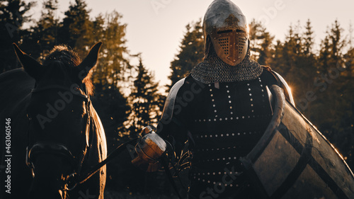 Medieval Knight Warrior Leading His Horse into the Battle to Fight. Portrait of Hero in Plated Armor, Shield, Helmet, Sword and Thoroughbred Stallion. Cinematic Light, War, Smoke, Invasion, Conquest © Gorodenkoff