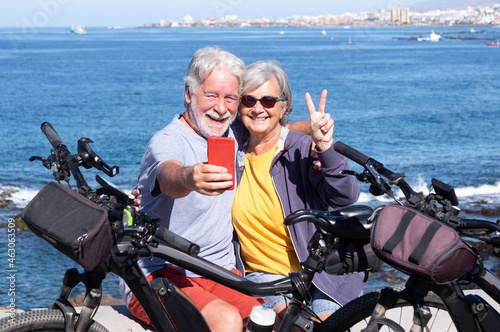 A joyful senior couple enjoying sea excursion with their bicycles, horizon on the sea. Active beautiful retirees enjoying healthy lifestyle using mobile phone for a selfie