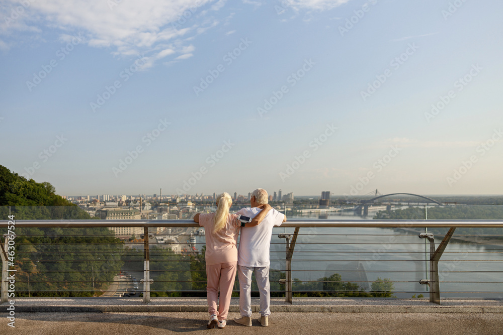Mature woman hugs man in sportsweat leaning onto railing on contemporary footbridge