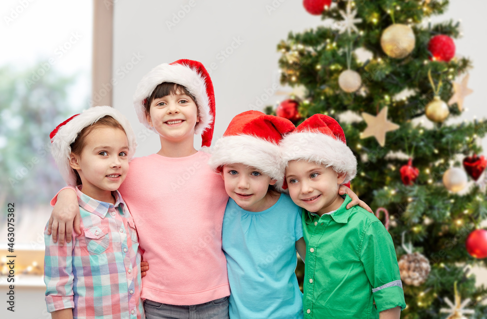 winter holidays, friendship and people concept - group of happy smiling little children in santa hats hugging at home over christmas tree background winter holidays, friendship and people concept - group of happy smiling little children in santa hats hugging at home over christmas tree background