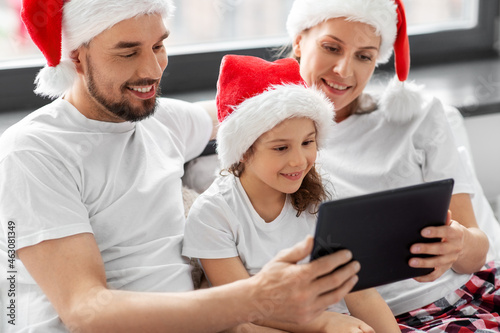 family, winter holidays and people concept - happy mother, father and little daughter with tablet pc computer in bed at home on christmas