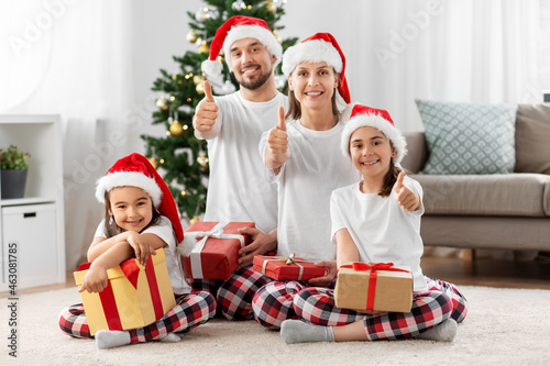 family, winter holidays and people concept - happy mother, father and two daughters with gifts sitting on floor at home and showing thumbs up