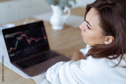 Young woman at home in the kitchen in a white hoodie with a laptop, graph on the screen, upset, unhappy, cryptocurrency falls down