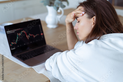 Young woman at home in the kitchen in a white hoodie with a laptop, graph on the screen, upset, unhappy, cryptocurrency falls down photo
