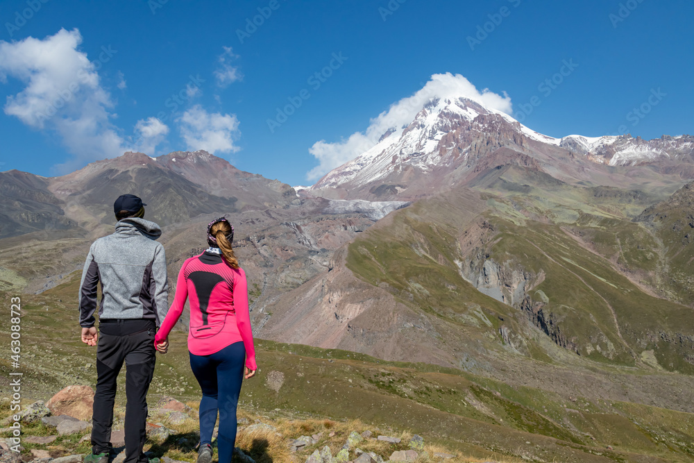 A couple enjoying the cloudless view on Mount Kazbeg in Caucasus, Georgia. There slopes are barren and stony below the snow-capped peak and the Gergeti Glacier. Tranquillity. Natural remedy. Adventure