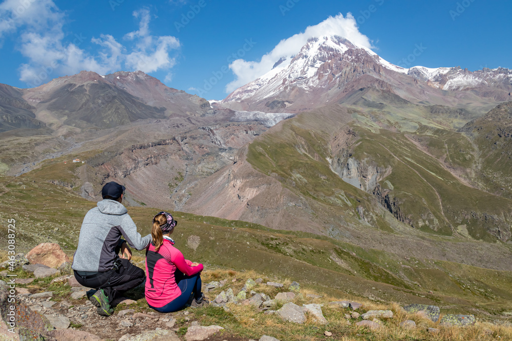 A couple enjoying the cloudless view on Mount Kazbeg in Caucasus, Georgia. There slopes are barren and stony below the snow-capped peak and the Gergeti Glacier. Tranquillity. Natural remedy. Adventure