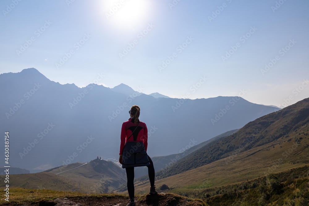 A woman enjoying the view on Caucasus mountains in Georgia. High mountain chains in front. There slopes are overgrown with green grass. Cloudless sky. Exploring new places