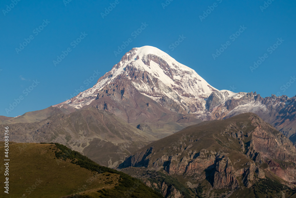 A cloudless view on Mount Kazbeg in Caucasus, Georgia. There slopes are barren and stony below the snow-capped peak and the Gergeti Glacier. Tranquillity. Natural remedy. Massive glacier foot A cloudless view on Mount Kazbeg in Caucasus, Georgia. There slopes are barren and stony below the snow-capped peak and the Gergeti Glacier. Tranquillity. Natural remedy. Massive glacier foot