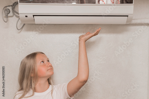 Young cute girl teenager blonde European appearance brings his hand to the air conditioner on the wall to check the heat and cold, the temperature in the room
