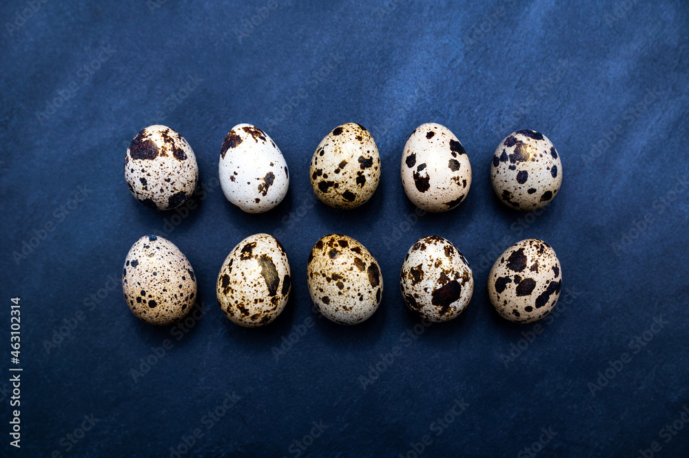 Ten typical patterned quail eggs (with a little gold dust) in two rows on a dark slate serving plate. Table top view of a kitchen, restaurant, or cooking scene.