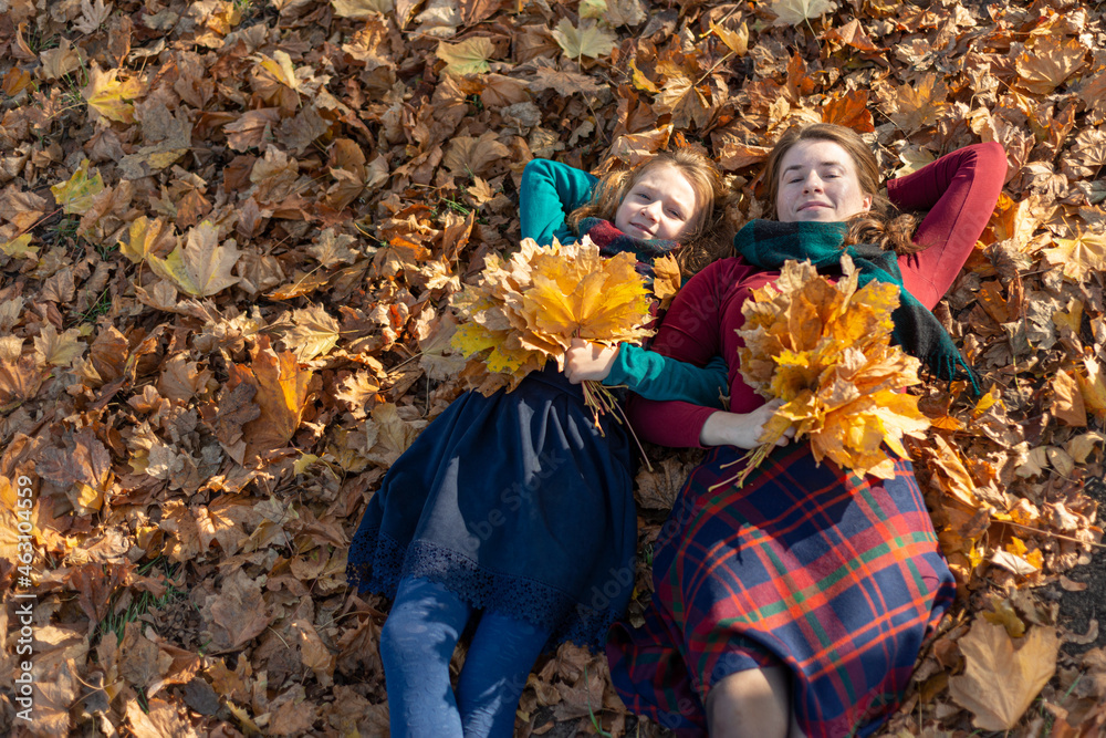 Mom and daughter lie on autumn leaves in the park and hold a bouquet of yellow leaves in their hand.