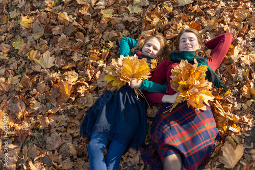 Mom and daughter lie on autumn leaves in the park and hold a bouquet of yellow leaves in their hand.