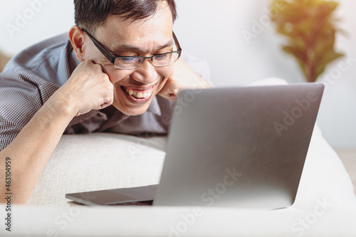 A cheerful Asian man glances at his laptop computer during a webcam video chat conference call in his living room, on the couch
