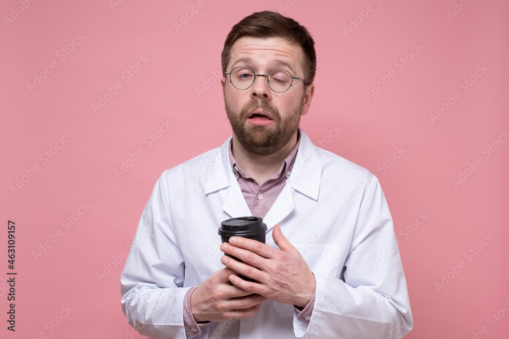 Doctor in a white coat and glasses is very tired and looks sleepy, he holds a paper cup of coffee in hands and tries to open his eyes. Doctor in a white coat and glasses is very tired and looks sleepy, he holds a paper cup of coffee in hands and tries to open his eyes.