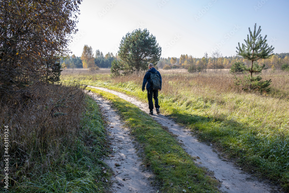 Obraz premium A tourist walks along a country road early in the morning. A man with a backpack on the background of a field illuminated by sunlight.