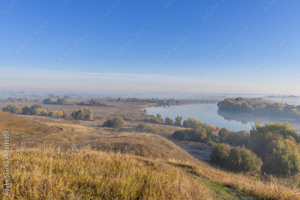Autumn landscape in the early morning overlooking the river. A wide river and endless expanses of fields. Yellow leaves on trees and bushes are illuminated by the rays of the rising sun. Autumn landscape in the early morning overlooking the river. A wide river and endless expanses of fields. Yellow leaves on trees and bushes are illuminated by the rays of the rising sun.