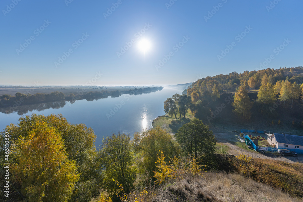 Obraz premium Autumn landscape in the early morning overlooking the river. A wide river and endless expanses of fields. Yellow leaves on trees and bushes are illuminated by the rays of the rising sun.