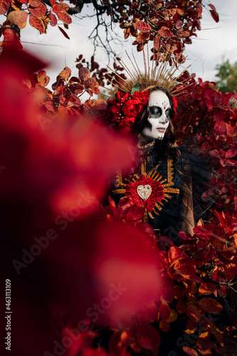 Woman with sugar skull makeup and red roses dressed as Santa Muerte is against background of autumn forest. © Stanislav
