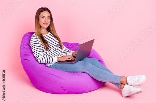 Photo of pretty shiny young woman dressed striped pullover sitting bean bag working modern device smiling isolated pink color background