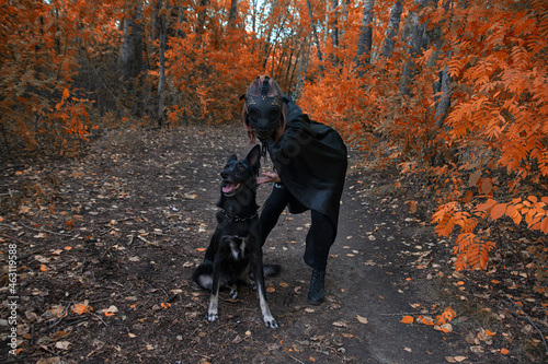 a girl in a bdsm costume and a black mask and with a dog in red forest.an an idea for Halloween.
