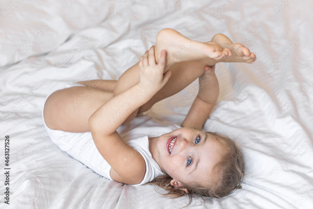 Toddler smiling baby girl in a white bodysuit lying on bed at home looking at the camera