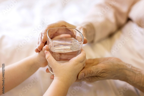 Small child gives a glass of water to an elderly person to a grandmother in the bed at home. Close-up of a hand with a glass. The concept of caring for adults, support, love, assistance